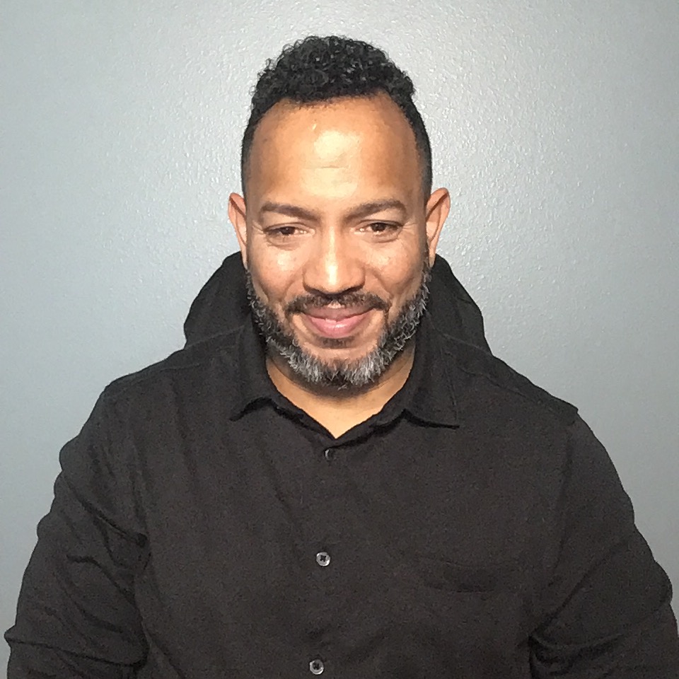 A man with a short, brown beard with tufts of grey. His short hair is slightly longer on the top, and he glances slightly downward. He's wearing a black button-op cardigan with a hood draped backward.