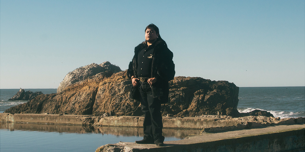 Against a blue sky, a person stands with short black hair by a body of water.