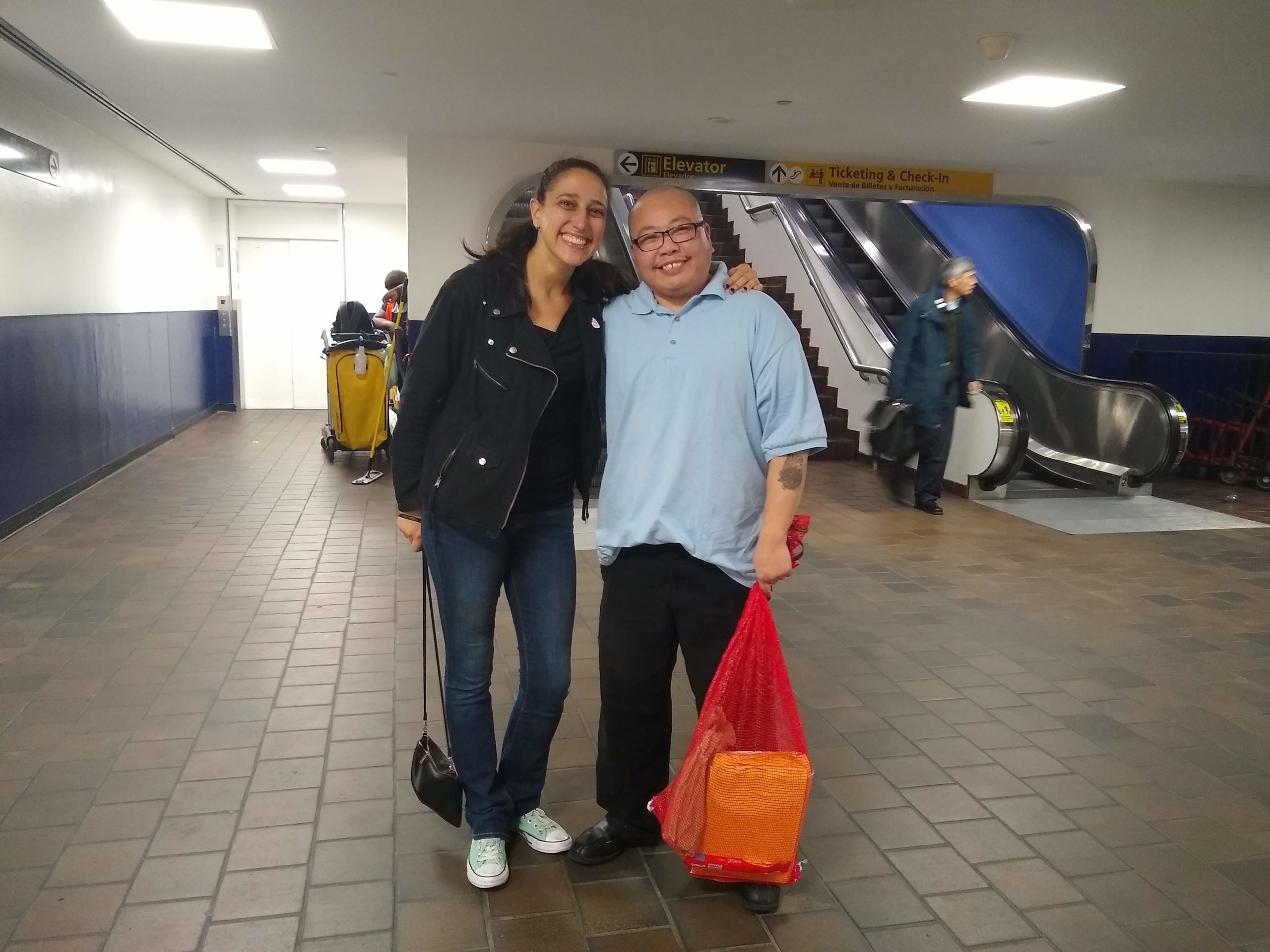 Chin and Lynly smiling and hugging, facing the camera. Chin is holding a red mesh bag with his belongings.
