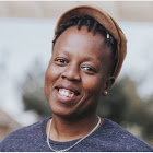 A Black person with short dark hair, grinning wide with their head held at an angle. They wear a brown short-brimmed cap, a blue t-shirt, and a silver metal necklace.
