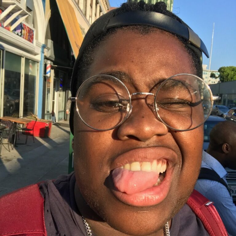 A black person who is playfully sticking their tongue out, they are wearing big glasses and a headband.