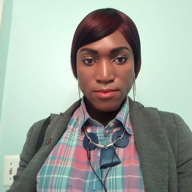 A black person with their hair neatly to the back . they are wearing a grey sweater, earing that dangle and a necklace, with a buttoned up shirt.