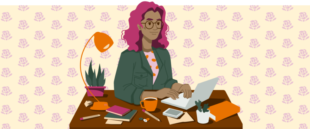 A brown person with wavy pink hair sits at a desk, typing on a small silver laptop. They’re wearing a green jacket with a pocket on the chest, and a pink shirt with orange polka dots. Various items line the desk, including several notebooks, a coffee mug, pencils, buttons, a lamp, and a plant. Behind them is yellow wallpaper with rows of pink decorative roses.