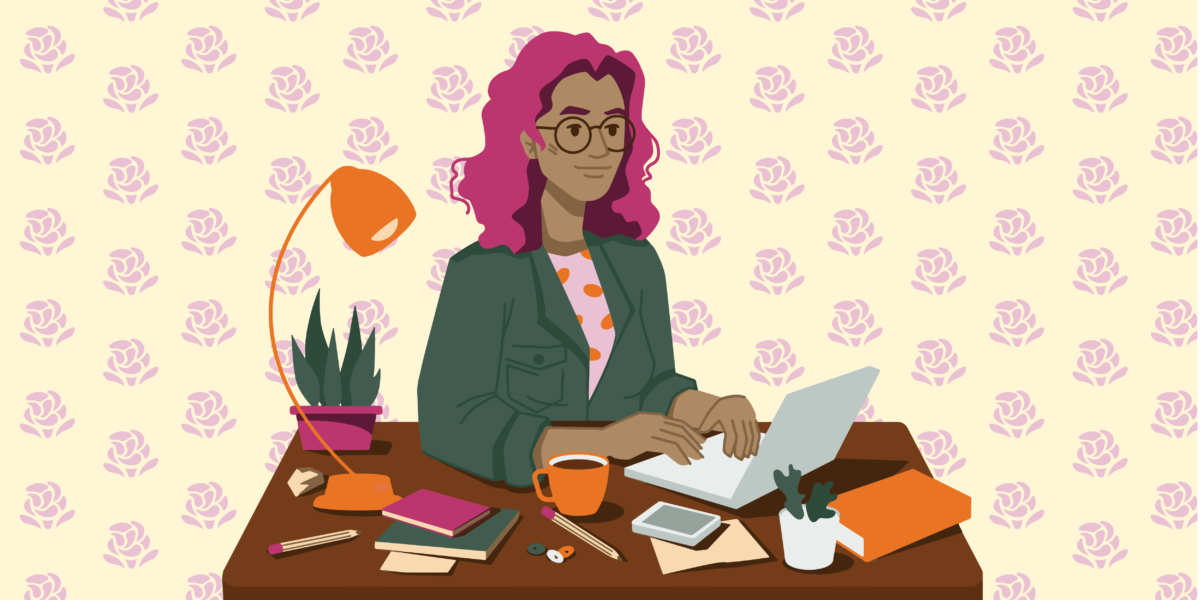 A brown person with wavy pink hair sits at a desk, typing on a small silver laptop. They’re wearing a green jacket with a pocket on the chest, and a pink shirt with orange polka dots. Various items line the desk, including several notebooks, a coffee mug, pencils, buttons, a lamp, and a plant. Behind them is yellow wallpaper with rows of pink decorative roses.