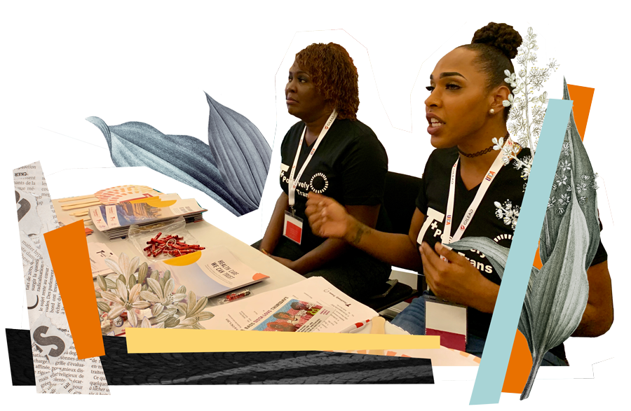 Two black feminine people, sitting at a table with boards behind them saying: "T+ Positively". on the table infront of them lay brochures and pamphlets.